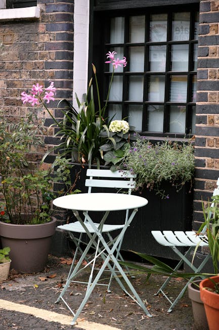 A cozy urban garden corner featuring a small white metal bistro table and matching chairs placed on a paved patio area outside a brick building. Various potted plants with lush green leaves and vibran